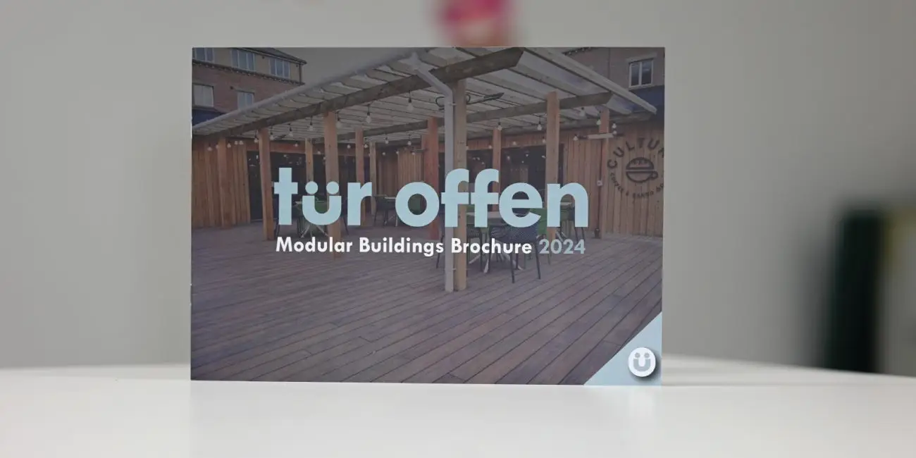 [A brightly colored brochure laid open on a wooden table, showcasing vibrant images of modular buildings on the left side, with detailed text descriptions on the right. The brochure highlights the benefits of modular construction in terms of efficiency, sustainability, and design flexibility. The Tür Offen logo is prominently displayed at the top.