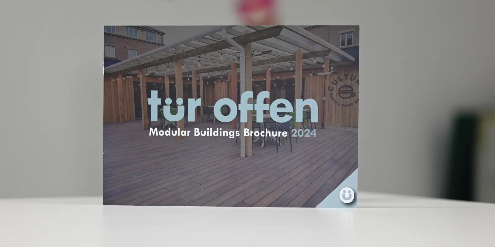 [A brightly colored brochure laid open on a wooden table, showcasing vibrant images of modular buildings on the left side, with detailed text descriptions on the right. The brochure highlights the benefits of modular construction in terms of efficiency, sustainability, and design flexibility. The Tür Offen logo is prominently displayed at the top.